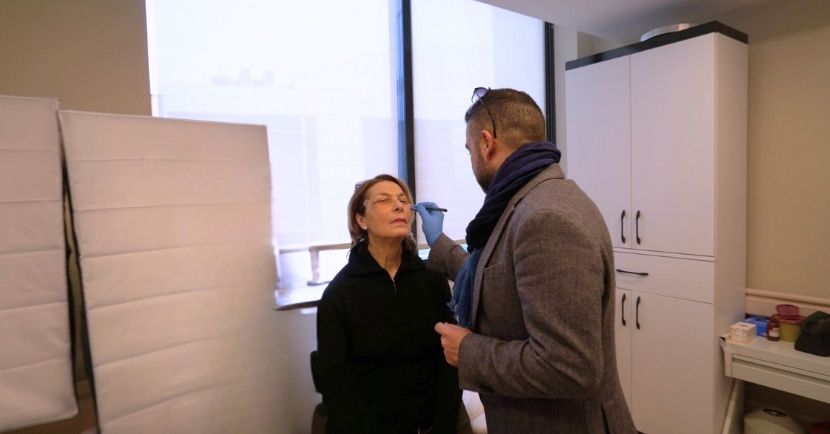A person in a black jacket sits with eyes closed while another in a gray blazer examines their face, possibly consulting for a facelift. Theyre in a room with a large window and white cabinets.